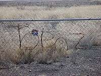 Photograph of the word "Topaz" written in barbed wire strung on a chain-link fence, at the Central Utah Relocation Center in 2006.