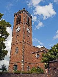 A two-storey brick church with a tall slim tower. It has clock faces and a battlemented parapet