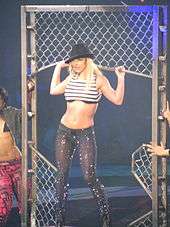 A blond female performer. She is standing inside a moving jungle gym-like metal structure. She is being carried by two people, who are halfway out of the picture. Her hands are grabbing the structure. She is looking to the left side of the picture. She is wearing a black hat leaning down to the left, a striped top exposing her midriff and sparkly black pants.