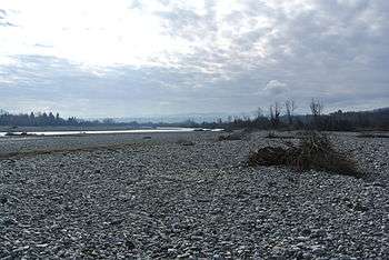 Photo shows a river bordered by an immense bed of white stones. There are mountains in the distance.