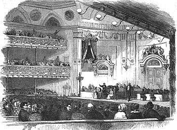 The interior of an elegant concert hall, an elegant audience, and on the stage, many men participating in the ceremony.
