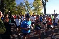 Three women, two smiling, and a man with his hand pointing into the air leading a large group of runners past Lake Calhoun and some observers