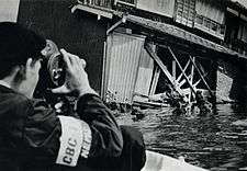 Black and white image of a cameraman in flood waters taking an image of a building's damaged façade.