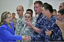 Clinton greeting U.S. military personnel at Andersen Air Force Base in Guam. The personnel are wearing uniforms and standing side-by-side.
