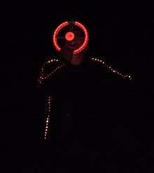 Close up, the dark-clothed singing man has lights embedded in his jacket sleeves and side, and is singing into a microphone with an illuminated circular structure around it, somewhat like a steering wheel, that he is holding with one hand.