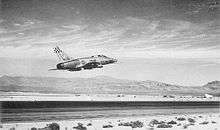 Rear quarter view of a single engine jet fighter taking off from a runway located in a desert. The bare metal finish jet has a checkerboard unit insignia on its vertical tail. The landing gear are raised. The horizon is hilly.