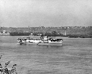 A World War II-era aircraft carrier at anchor. Several aircraft are on her flight deck and land and buildings are visible in the background.