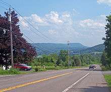A two-lane road descending left to right across the bottom half of the image, with telephone poles along its left side, to a right turn. In the distance is a valley that extends to the rear of the field of view.