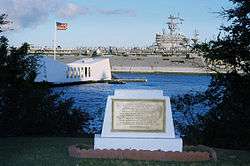 A golden plaque on stone on the shore of Ford Island, with a white memorial bridge floating over the USS Arizona