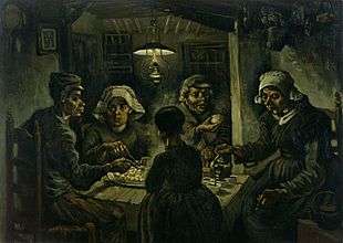 A group of five sit around a small wooden table with a large platter of food, while one person pours drinks from a kettle in a dark room with an overhead lantern.