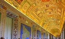 Photo of a long wide corridor filled with a crowd of people in casual dress. The ceiling is arched and is elaborately decorated with gilt stucco and small brightly coloured pictures. The walls have frescoes of large maps, each of which has a brilliant blue background.
