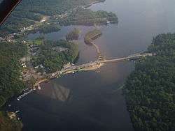 A large lake is surrounded by land partially or wholly covered in forests. A road crosses the lake via bridge and intersects another that crosses a small bay by way of a narrow strip of land. At lower left is a community composed mostly of houses. The highway carried by the bridge across the lake serves the community and intersects another highway at upper left.