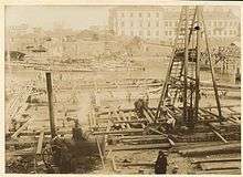 Sepia photograph of construction works in Vilna's borough of Zwierzyniec. The river is filled with wooden pontoons and elements of wooden scaffolding to be erected, in the foreground on the near bank of the river a steam engine is visible on the left, on the right a group of workers are operating a tall wooden pile driver. On the far bank a similar pile driver is visible, as well as some townhouses.