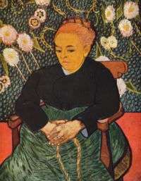 An elderly well-dressed woman sits facing to her right (the viewer's left). She has her hands clasped together on her lap, and she is dressed in a dark top and green dress in front of a vivid flower wallpaper background.
