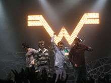 Four men stand in front of an audience; two of them raise their arms. Behind them is an illuminated sign in the shape of the letter "W".