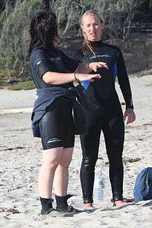 Two women in wetsuits standing on a beach. Left is wearing a shortie suit, also called a "spring suit", which has short legs and sleeves, with boots. Right is wearing a full length one piece suit with back zipper, also called a "steamer".
