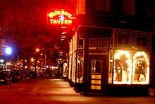 On the corner of a block is a building with large glass fronts on both sides; a sign displaying the tavern's name shines brightly above in red neon.