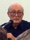 An elderly man wearing glasses with a microphone in front of him on the left side.