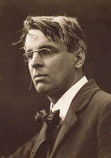 A posed black and white photograph of Yeats. He is wearing smart clothes and spectacles, while his hair looks a bit tousled