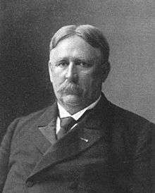 Head and shoulders of a white man with hair parted in the center and a mustache, wearing a dark suit and tie.