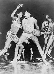 A basketball player, wearing a light-colored jersey with the number "13", is holding a basketball in front of another basketball player who is wearing a dark-colored jersey with the number "6".