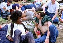 A color photograph showing people from the 1969 Woodstock Festival sitting on grass, in the foreground a back and a white male look at each other