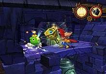 A character in brightly colored attire rings an oversized, gold and blue bell as he stands on a dark, stone platform. Directly in front of him is a green-colored cartoon bomb with a lit fuse. Below the platform is larger bell not completely in view. The upper righthand corner of the image shows two circular icons, the first showing a skull-and-crossbones with rabbit ears and the second showing a movie camera.